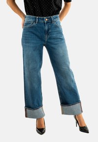Jeans blu a vita alta con vestibilità rilassata, orli risvoltati e dettagli in contrasto sulle cuciture interne, abbinati a tacchi alti neri.