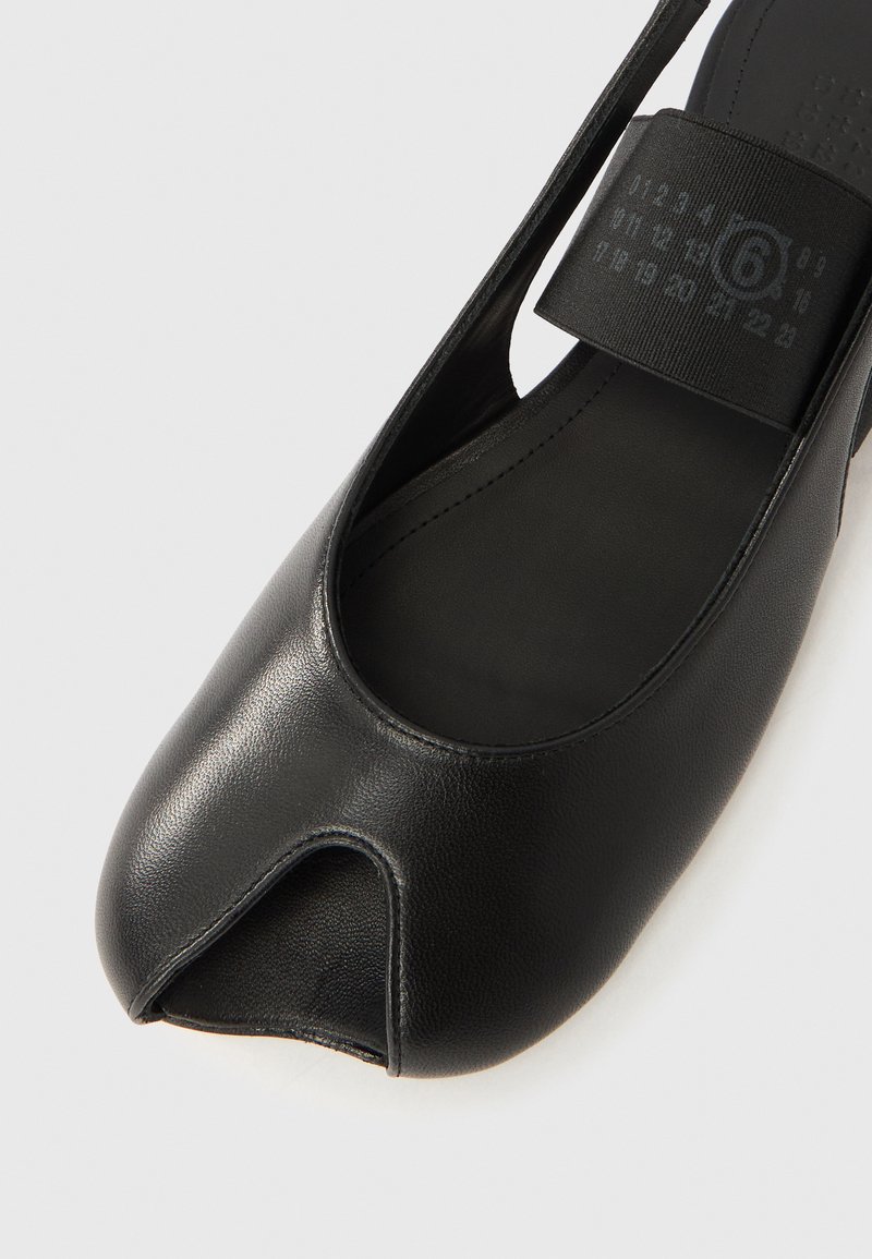 Sort læder flad sko med delt tå, bred elastisk rem og synlige størrelsesmarkeringer på remmens inderside, vist mod en hvid baggrund.