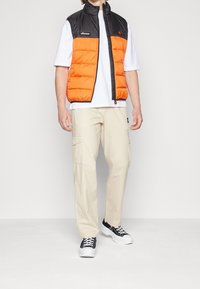 Doudoune sans manches avec panneaux orange et noir, tissu léger, fermeture éclair frontale, portée avec un t-shirt blanc et un pantalon cargo beige, avec poches.