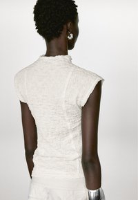 Witte textuurtop met korte mouwen en een hoge kraag, met een gekreukt ontwerp. De rug van het model is zichtbaar, met een metalen armband.