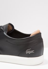 Black leather sneaker with a white rubber sole, featuring a beige accent at the heel and a silver crocodile logo on the side.