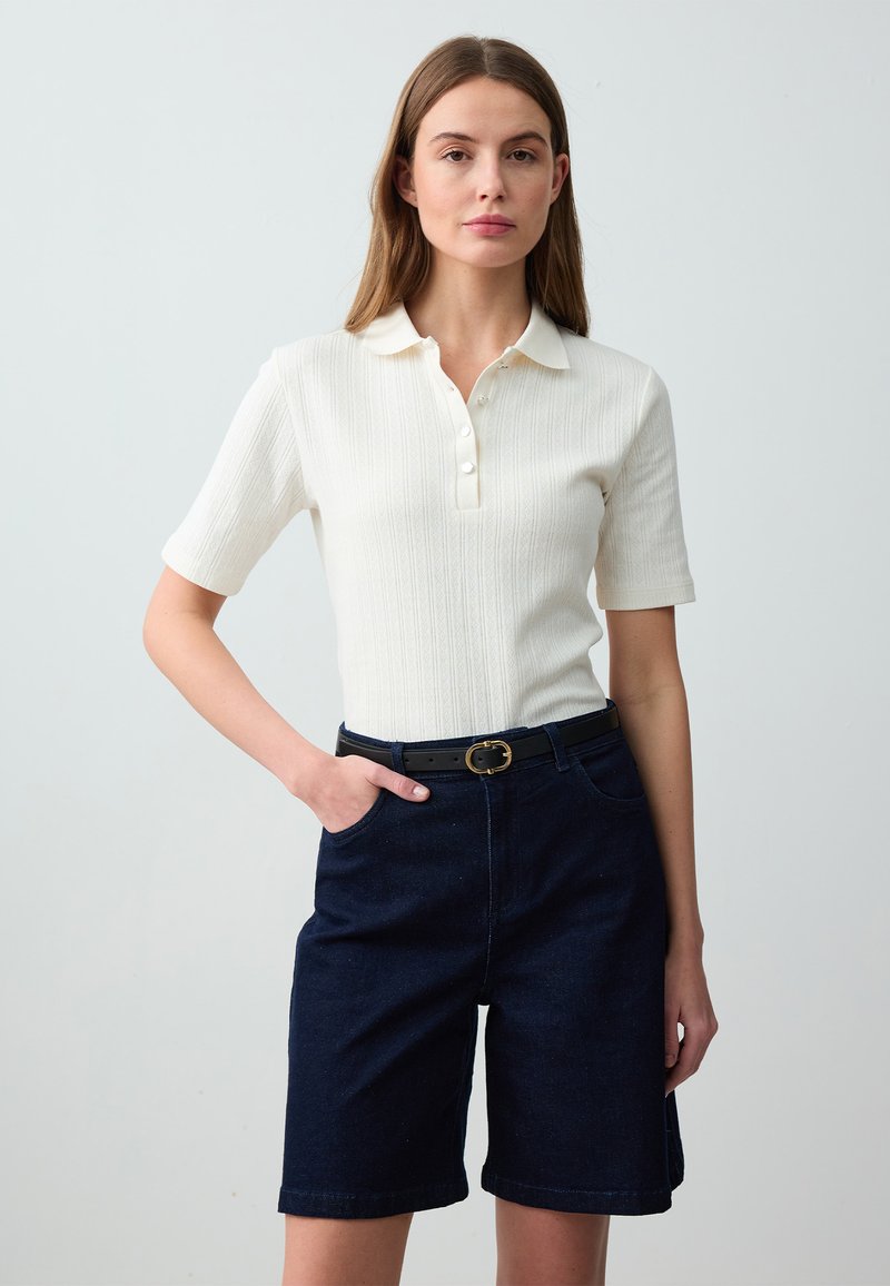 Polo bianco a coste con maniche corte e patta dei bottoni, abbinato a pantaloni corti in denim blu navale e una cintura nera con fibbia dorata.