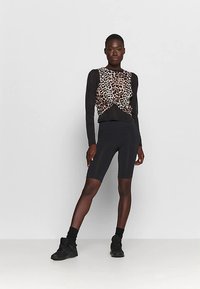 Zwart lange mouwen top met een luipaardprint aan de voorkant, gecombineerd met zwarte fietsbroek en zwarte sneakers. Model staat voor een neutrale achtergrond.