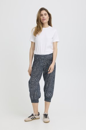 Žena stojaca, oblečená v bielom tričku s krátkym rukávom, tmavomodrých vzorovaných skrátených nohaviciach a béžových teniskách s čiernymi pruhmi, na jednoduchom svetlom pozadí.