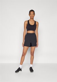 Svart sportbh och svarta träningsshorts med elastisk midja, tillsammans med svarta skor och vita strumpor, på en enfärgad bakgrund.