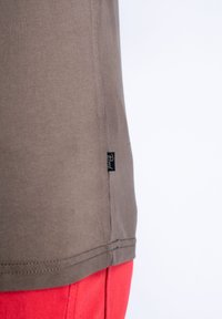 Brown cotton t-shirt with a straight hem, side seam label, and visible stitching. Paired with red fabric shorts.