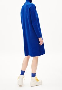 Person klädd i en knälång, långärmad royalblå klänning med matchande strumpor och vita klumpiga stövlar med gula snören, stående framför en vit bakgrund.