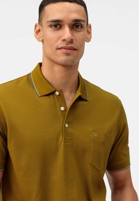 Olijfgroene polo met drie witte knopen, een borstzak en blauwe kraagaccenten. Gelaagd stof met een aansluitend ontwerp.