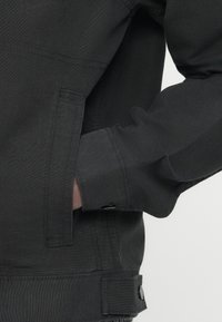 Μαύρο τζιν μπουφάν με μανσέτα με κουμπιά, εμφανή ραφές και πλαϊνή τσέπη. Το ύφασμα έχει ομαλή υφή με δομημένο σχεδιασμό.