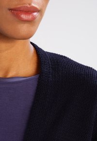 Námornícky pletený cardigan s texturovaným vzorem, navržený s otevřeným předním dílem a měkkým fialovým topem pod ním, který zdůrazňuje hladký výstřih.