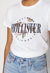 White t-shirt with "Hollister Southern California" text, floral design, and orange butterflies, worn with light blue denim jeans.