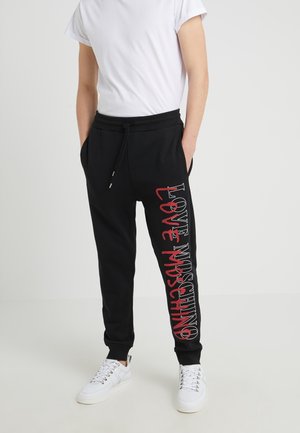 Osoba nosící černé tepláky s nápisem "LOVE MOSCHINO" v bílé a červené barvě na jedné noze, doplněné o jednoduché bílé tričko a bílé tenisky.