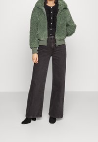 Grön fakepälsjacka med dragkedja och ribbade ärmslut och nederkant, ihopparad med högmidjade svarta vida jeans. Svart topp med volangdetaljer.
