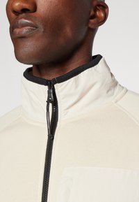 Giacca zip-up color crema con colletto alto, cerniera nera e tessuto a trama. Presenta una tasca sul petto e dettagli di cucitura a contrasto.