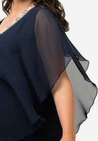 Dunkelbluse mit einem transparenten, fließenden Cape-Ärmel und einem verzierten Ausschnitt mit perlenähnlichen Akzenten, hergestellt aus leichtem Stoff.