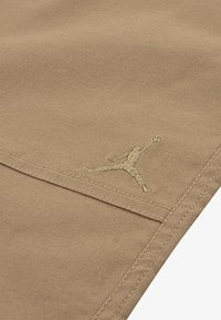 Tan katoenstof met een gladde textuur heeft een gouden geborduurd Jumpman-logo. Zichtbare stikseldetails langs de randen.