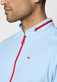 Chaqueta azul claro con un alto cuello, que presenta una cremallera roja y un pequeño emblema bordado con tres hojas. Textura de tela suave.