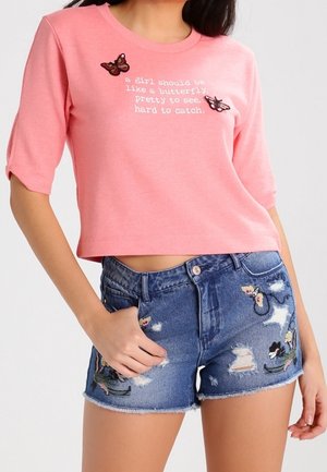 Vrouw draagt een roze cropped shirt met vlinderapplicaties en witte tekst, gecombineerd met gescheurde blauwe spijkershorts met bloemenborduursel.