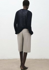 Pulover tricotat semitransparent de culoare navy, cu mâneci lungi, asociat cu pantaloni scurți bej, ajustați, și pantofi negri fără toc, văzut din spate.