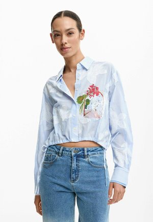 Nő világoskék, rövidített, gombos ingben, fehér virágmintákkal és piros virágmotívummal, magas derekú kék farmerrel párosítva.