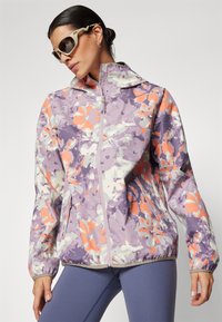Chaqueta ligera con capucha en un patrón floral de color púrpura, naranja y blanco. Presenta un cierre frontal y puños elásticos con un corte ajustado.