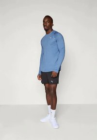 Hosszú ujjú kék sportpóló, kerek nyakkivágással, fekete rövidnadrággal párosítva. Mindkettőn Puma logó található. A modellen fehér zoknik és cipők vannak.