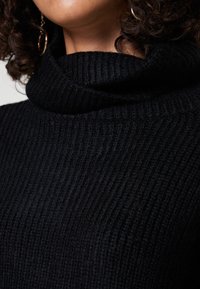 Vrouw met krullend haar, gekleed in gouden oorbellen en een zwart geribde turtleneck trui.