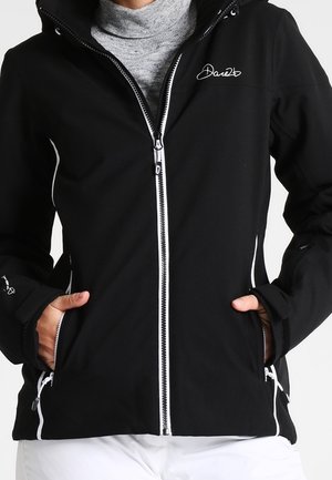 Zwarte jas met ritssluiting, witte bies en logo op de borst, gedragen over een grijze coltrui, met handen in de voorzakken.