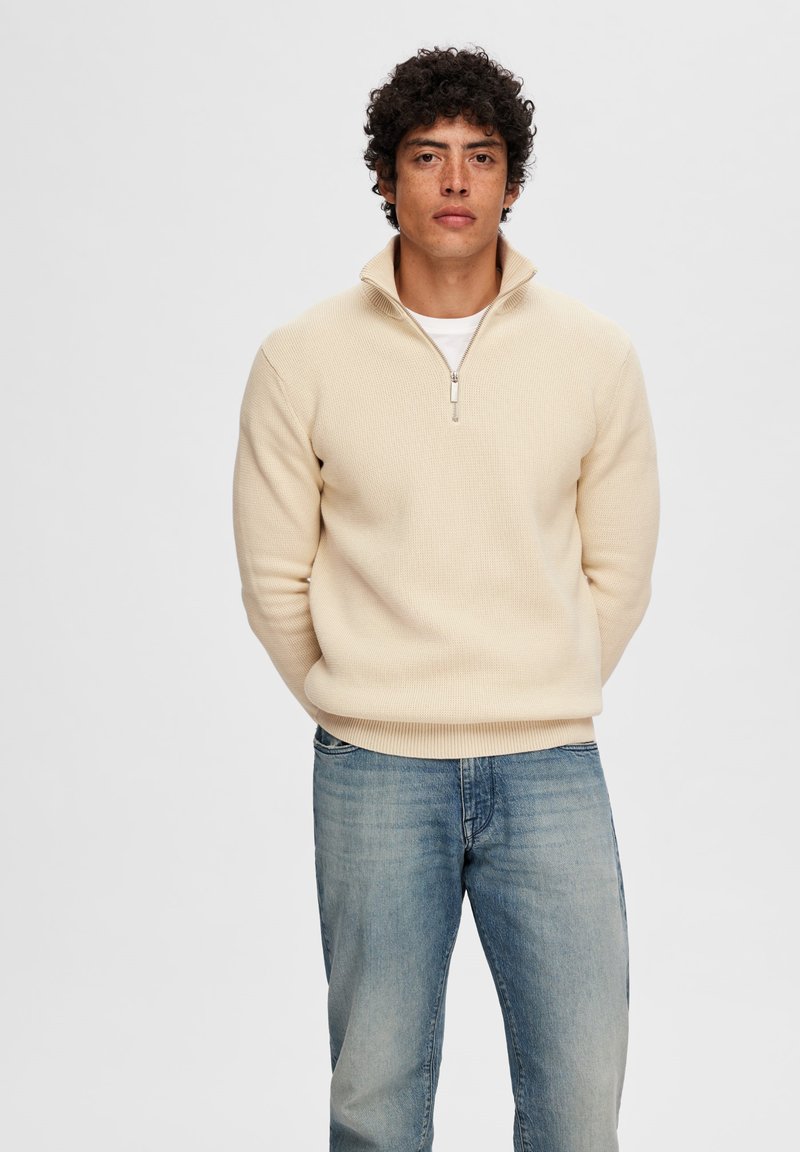 Cremfarbener Pullover-Sweatshirt mit Reißverschlusskragen, gerippter Textur und weichem Stoff. Trägt man über einem weißen T-Shirt und kombiniert mit hellblauen Jeans.