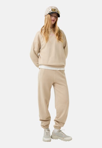 Beige oversized sweatshirt og jogger-sæt i blødt stof, med elastiske manchetter og en lagdelt hvid skjorte under. Lyse sneakers.
