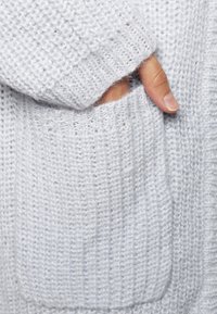 Hellgrauer Strickcardigan mit strukturiertem Muster, ausgestattet mit einer sichtbaren Seitentasche und gerippten Bündchen. Weicher, entspannter Schnitt.