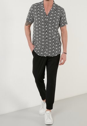 Mand iført sort og hvid skjorte med geometrisk mønster og korte ærmer, sorte joggingbukser, hvide sneakers, med den ene hånd i lommen.
