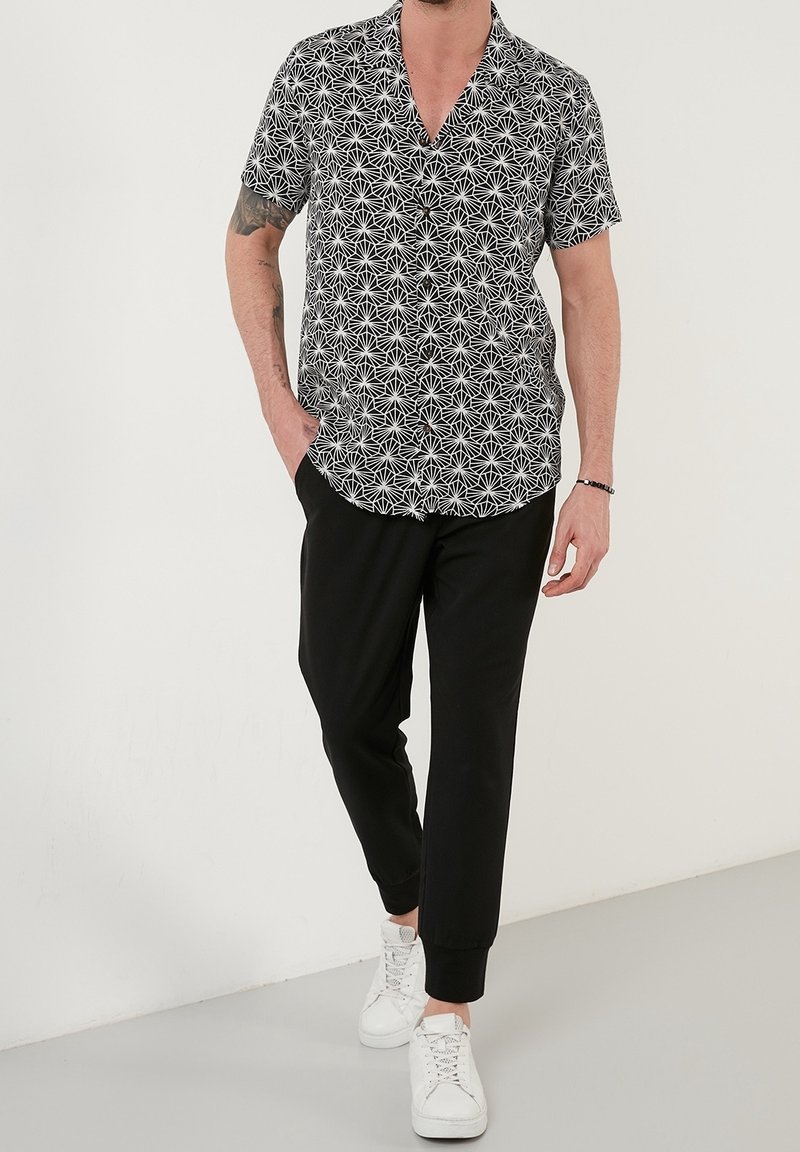 Homme portant une chemise à manches courtes à motif géométrique noir et blanc, pantalon de jogging noir, baskets blanches, avec une main dans la poche.