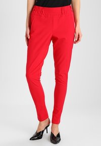 Pantalons rouges ajustés en tissu extensible. Dotés d'une taille élastique et de deux poches latérales. Portés avec des chaussures noires à bout pointu.