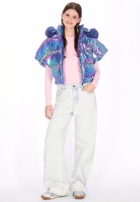 Perłowa, puchowa kamizelka z wysokim kołnierzem, ozdobiona dwoma niebieskimi pomponami na kapturze, noszona na różowym topie z długim rękawem i luźnych jasnych jeansach.