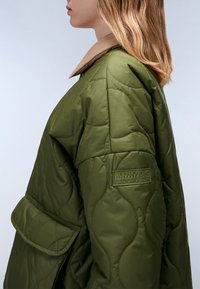 Giubbotto trapuntato verde oliva con colletto beige, caratterizzato da un logo a patch sulla manica e dettagli delle tasche laterali. Tessuto liscio, design di lunghezza media.