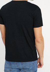 Hombre vistiendo una camiseta negra de manga corta lisa y jeans de mezclilla azules, de espaldas con la mano izquierda relajada a un lado.