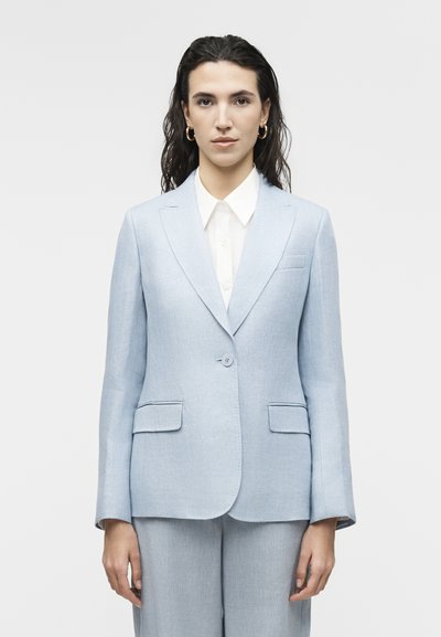 Femme aux cheveux bruns ondulés portant des boucles d'oreilles créoles en or, un blazer bleu clair à un bouton, une chemise blanche à col et un pantalon assorti.