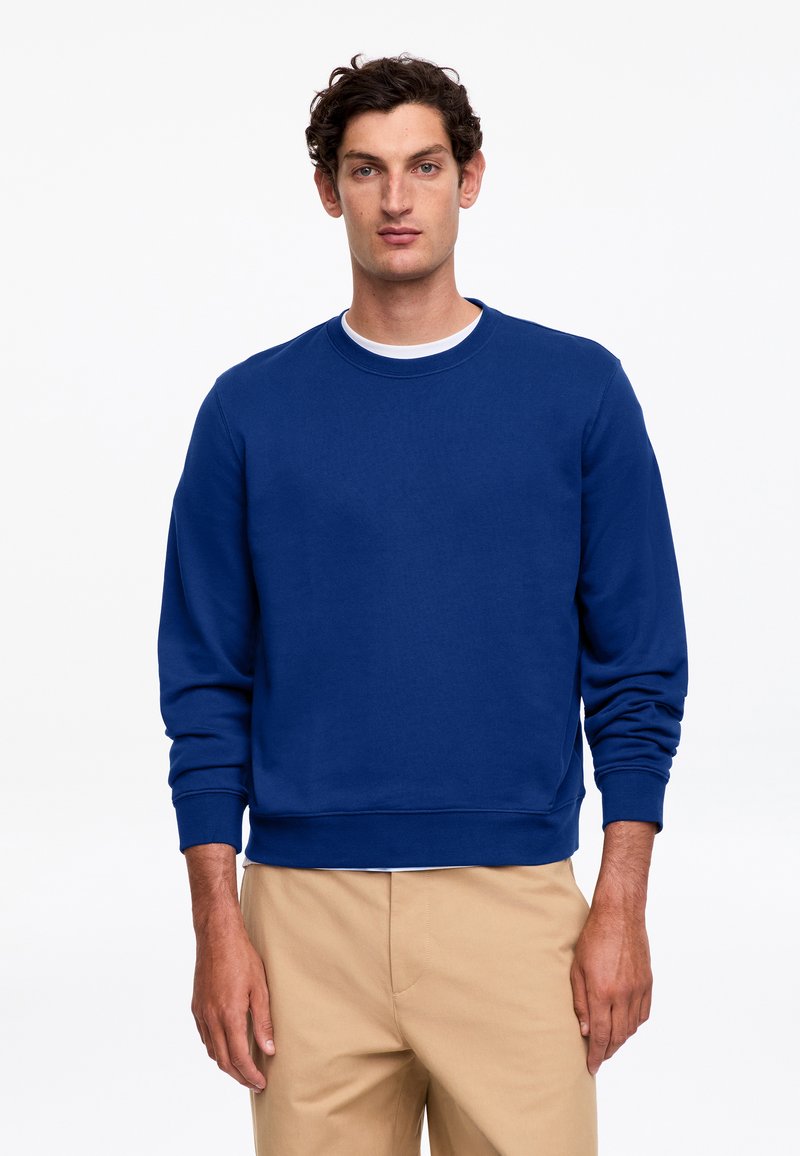 Homme aux cheveux courts et bouclés portant un sweat-shirt bleu uni à manches longues et un pantalon beige sur fond blanc.