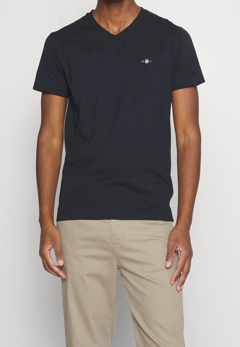 T-shirt noir en coton à col en V avec manches courtes, présentant un petit logo brodé sur la poitrine. Porté avec un pantalon beige.