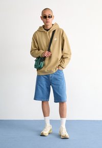 Hanorac bej, pantaloni scurți din denim albastru, șosete albe și adidași crem completează ținuta. Accesoriile includ o geantă verde tip crossbody și ochelari de soare galbeni.