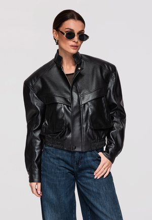 Frau trägt schwarze Lederjacke, dunkle Sonnenbrille, goldene Creolen und blaue Jeans, posiert mit einer Hand in der Tasche.