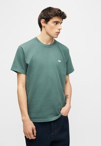 Νεαρός άνδρας με κοντά καστανά μαλλιά, φορώντας πράσινο t-shirt Obey και σκούρο τζιν, στέκεται με το ένα χέρι στην τσέπη, κοιτάζοντας προς το πλάι.