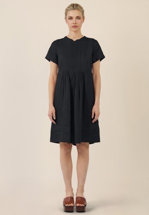 Kobieta o blond włosach w czarnej sukience do kolan z plisami, ubrana w brązowe sandały na platformie z odkrytymi palcami, stojąca na beżowym tle.