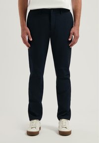 Marineblauwe broek met een getailleerde pasvorm, vlakke voorkant en riemlussen. Gedirigeerd met witte sneakers, die een gladde textuur en subtiele glans hebben.