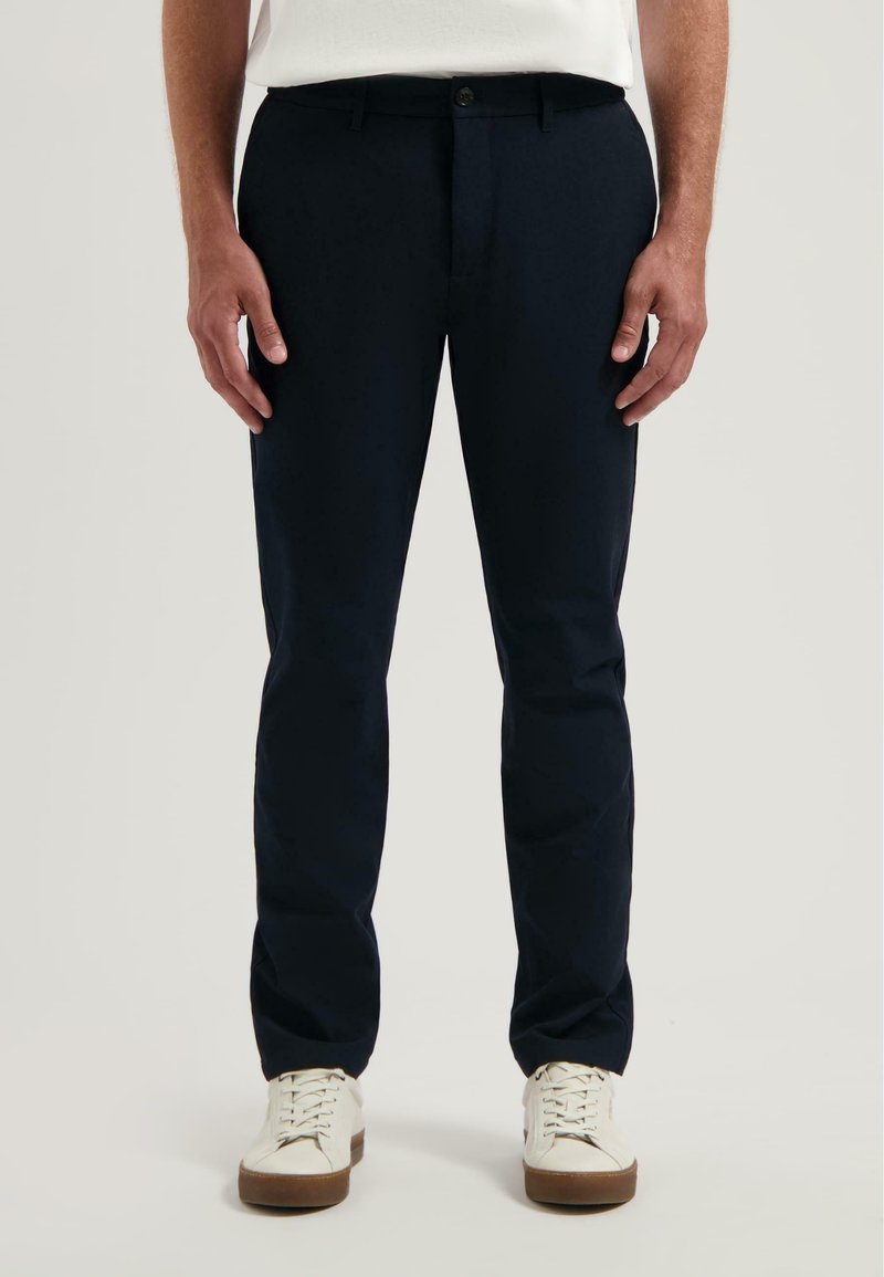 Marineblauwe broek met een getailleerde pasvorm, vlakke voorkant en riemlussen. Gedirigeerd met witte sneakers, die een gladde textuur en subtiele glans hebben.