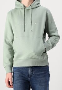 Světlé zelená mikina s kapucí z měkké látky, vybavená klokaní kapsou, stahovacími šňůrkami a bílým logem "JACK & JONES" přes prsa.