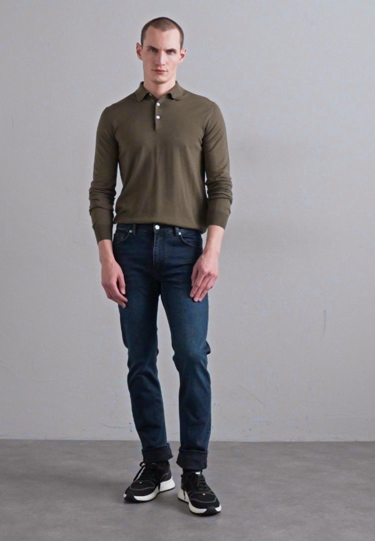 Polo à manches longues vert olive, coupe ajustée, associé à un jean bleu foncé, manches retroussées et chaussures de sport noires avec des accents blancs.