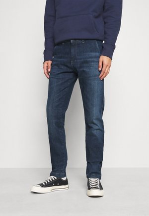 Uomo che indossa jeans blu scuro, sneaker nere e bianche e una felpa con cappuccio blu navy, in piedi contro uno sfondo grigio chiaro.