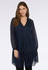 Granatowa sukienka o warstwowym kroju z lekkiego, przejrzystego materiału, z dekoltem w serek i długimi rękawami. Charakteryzuje się delikatnym połyskiem i elastycznymi mankietami.